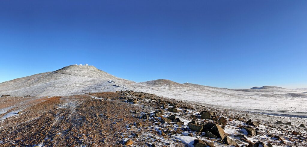 The Atacama Desert is one of the driest places in the world. Several factors contribute to its arid conditions. The magnificent Andes mountain range and the Chilean Coast Range block the clouds from the east and west, respectively. In addition, the cold offshore Humboldt Current in the Pacific Ocean, which creates a coastal inversion layer of cool air, hinders the formation of rain clouds. Moreover, a region of high pressure in the south-eastern Pacific Ocean creates circulating winds, forming an anticyclone, which also helps to keep the climate of the Atacama Desert dry. These arid conditions were a major factor for ESO in placing the Very Large Telescope (VLT) at Paranal, in the Atacama Desert. At the Paranal Observatory, located on the summit of Cerro Paranal, the precipitation levels are usually below ten millimetres per year, with the humidity often dropping below 10%. The observational conditions are excellent, with over 300 clear nights per year. The splendid conditions for astronomical observations in the Atacama Desert are only rarely disturbed by the weather. However, for perhaps a couple of days each year, snow pays a visit to the Atacama Desert. This picture shows a beautiful panoramic view of Cerro Paranal. The VLT is on the peak on the left, and the VISTA survey telescope is on a slightly lower peak, a short distance to the right. The blue sky shows that this is yet another clear sunny day. This time, though, something is different: a thin dusting of snow has transformed the desert landscape, producing an unusual view of rare beauty. This image was taken by ESO Photo Ambassador Stéphane Guisard on 1 August 2011. Links More about the VLT at Paranal ESO Photo Ambassadors